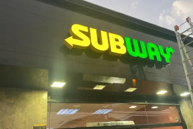 Letreiro em letras caixa iluminadas do Subway em amarelo e verde instalado em fachada preta de restaurante