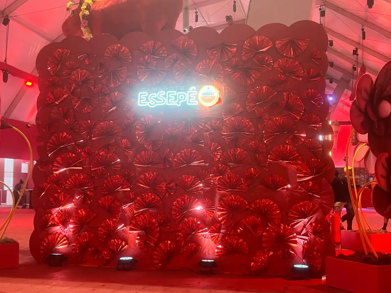 Letreiro neon Essepê sobre parede cenográfica de leques vermelhos